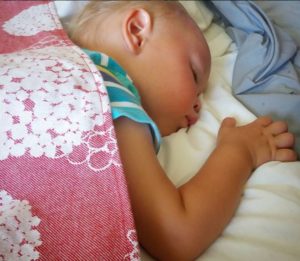 A sleeping toddler, on top of a bed, with a pink and white blanket draped over him.