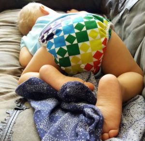 Toddler crashed out on the couch wearing a rainbow modern cloth nappy and a blur shirt.